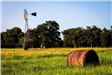 An image of a windmill and a round bale of hay resting in a field of tall green grass. 
